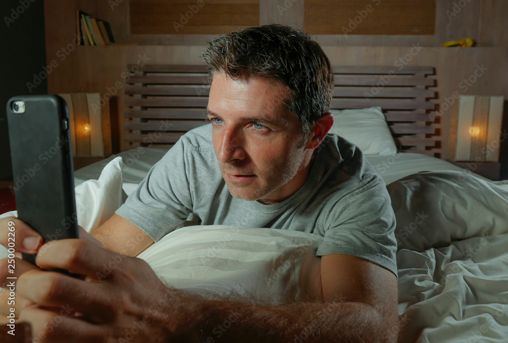 Obraz premium lifestyle home portrait of young attractive and relaxed man using internet social media app on mobile phone in his bedroom late at night lying on bed networking concentrated