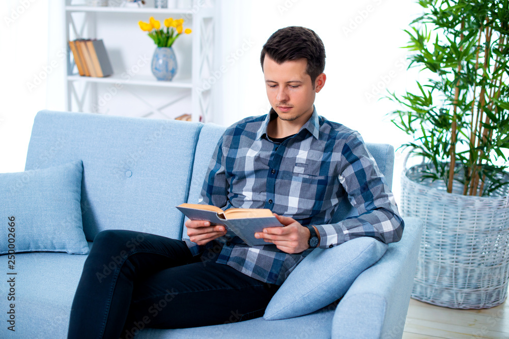 Handsome young man guy reading a book sitting on the couch in his living room