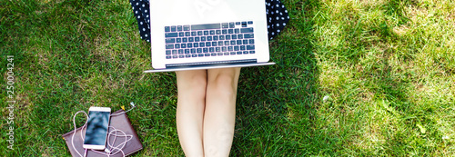 Top view of woman sitting in park on the green grass with laptop, notebook and smartphone, hands on keyboard. Computer screen mockup. Student studying outdoors. Copy space for text photo