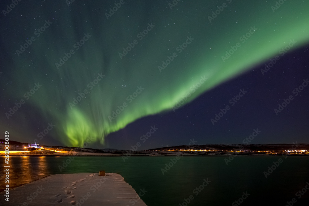 Drapery Aurora - Northern lights over a fjord with a snow covered pier in the foreground and a illuminated church in the background in Northern Norway