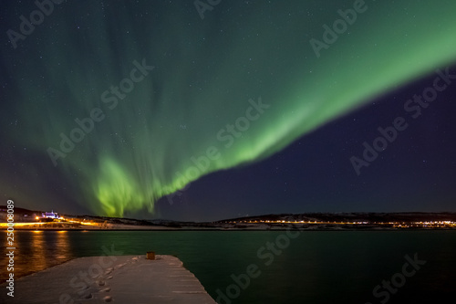 Drapery Aurora - Northern lights over a fjord with a snow covered pier in the foreground and a illuminated church in the background in Northern Norway