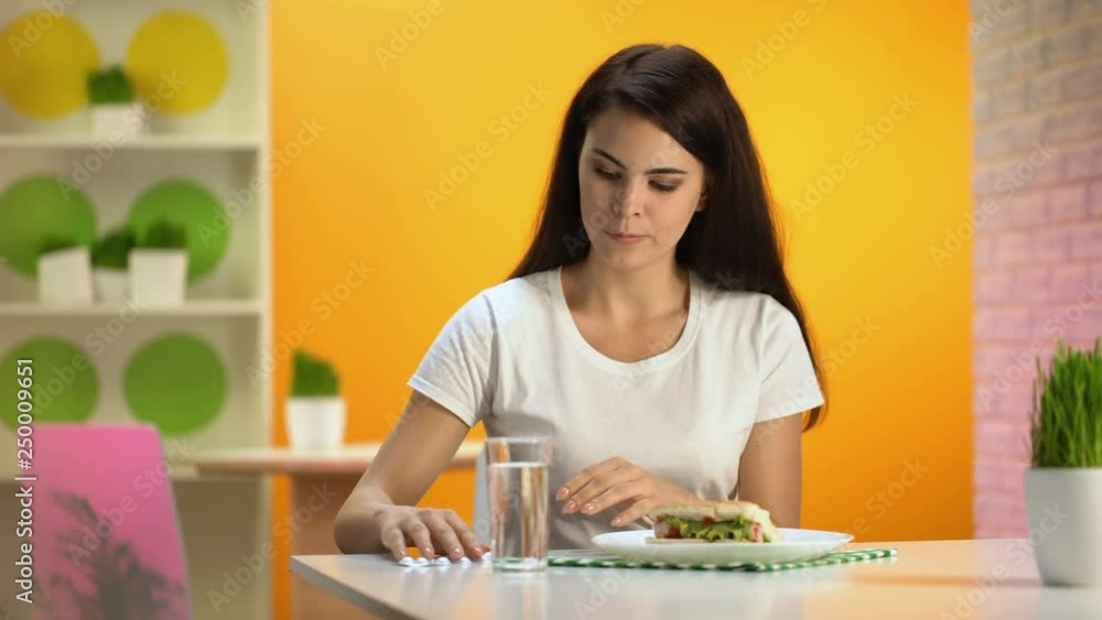 Pretty female taking pill with water sitting in fast food restaurant, overeating Pretty female taking pill with water sitting in fast food restaurant, overeating