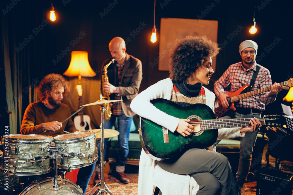 Fototapeta premium Mixed race woman singing and playing guitar while sitting on chair with legs crossed. In background drummer, saxophonist and bass guitarist.