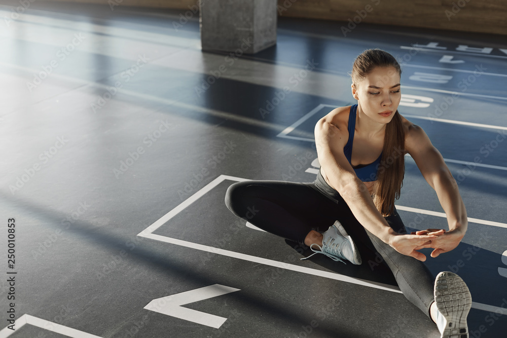 Focused woman athlete in sportswear sporty bra sitting gym floor stretching leg preparing muscles and biceps for working-out, pilates. Active young aerobics instructor practicing exercises Focused woman athlete in sportswear sporty bra sitting gym floor stretching leg preparing muscles and biceps for working-out, pilates. Active young aerobics instructor practicing exercises
