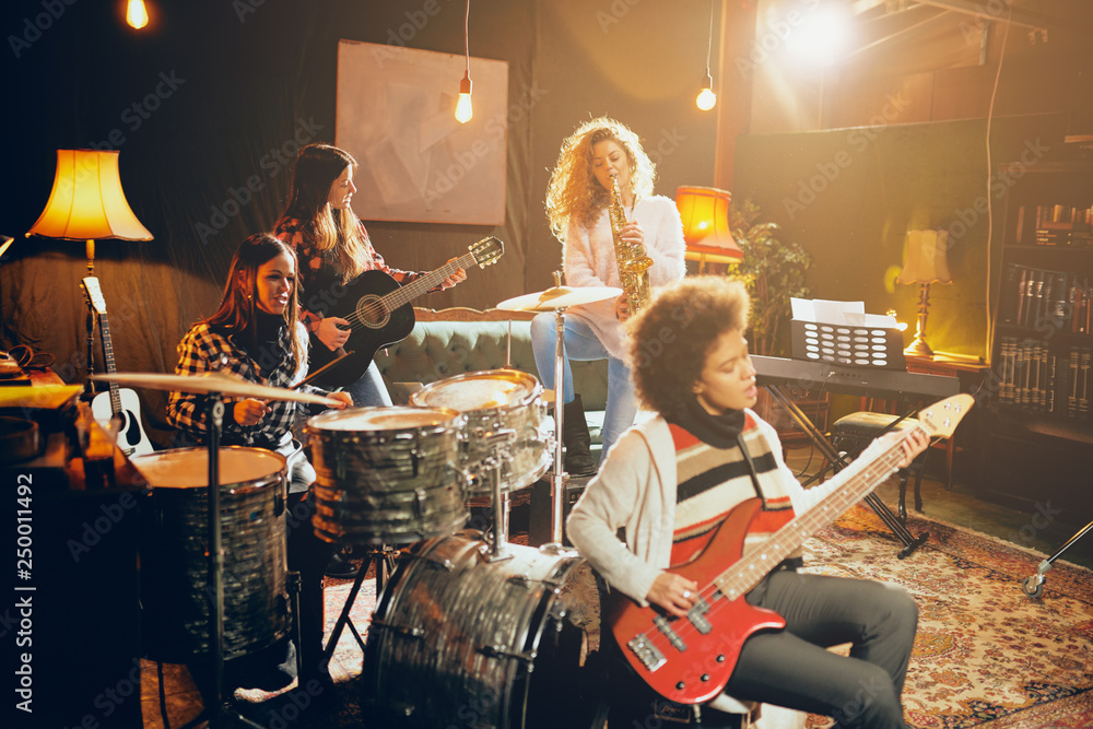 Naklejka premium Girls playing jazz music. In foreground one woman playing bass guitar and in background other three playing acoustic guitar, saxophone and drums. Home studio interior.