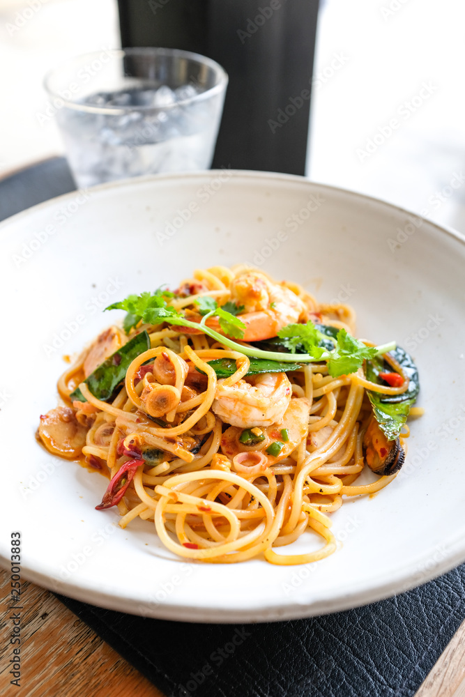 Spaghetti with spicy seafood Tom Yum sauce thai style in white ceramic plate on table in restaurant cafe. Modern fusion Thai food recipe.