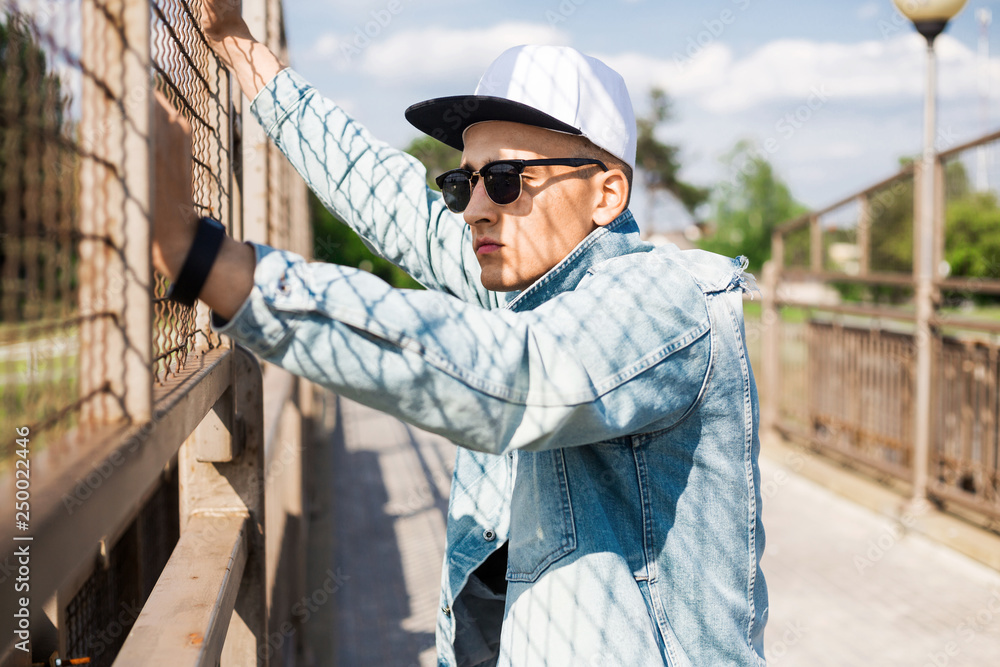 Stylish cute guy is standing on the bridge, wearing a denim jacket cap, listening to music through headphones free hands Stylish cute guy is standing on the bridge, wearing a denim jacket cap, listening to music through headphones free hands