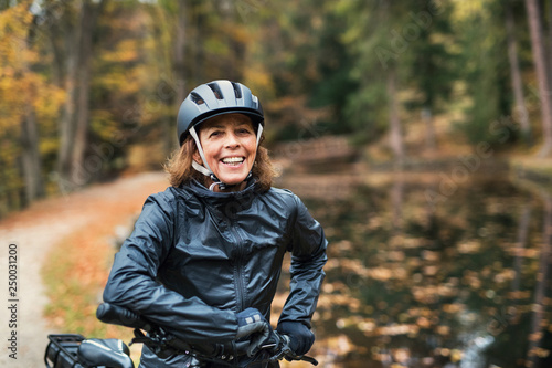 A senior woman with electrobike standing outdoors on a road in park in autumn.
