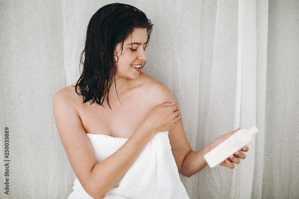 Young happy woman in white towel applying moisturizing cream on shoulder in bathroom. Skin and body care. Hand holding plastic bottle with lotion. Sexy woman relaxing, spa and wellness Young happy woman in white towel applying moisturizing cream on shoulder in bathroom. Skin and body care. Hand holding plastic bottle with lotion. Sexy woman relaxing, spa and wellness