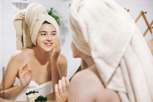 Skin Care concept. Young happy woman in towel making facial massage with organic face scrub and looking at mirror in stylish bathroom. Girl applying scrub cream, peeling and cleaning skin photo