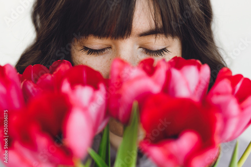 Beautiful brunette girl portrait with red tulips closeup on white background indoors, space for text. Stylish young woman  smelling tulips with closed eyes. Fresh aroma scent concept #250036075