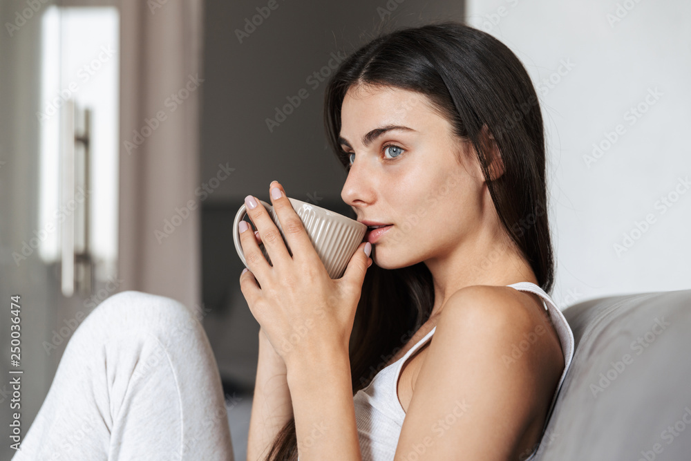 Pretty beautiful young woman sitting on sofa indoors at home drinking coffee or tea. Pretty beautiful young woman sitting on sofa indoors at home drinking coffee or tea.