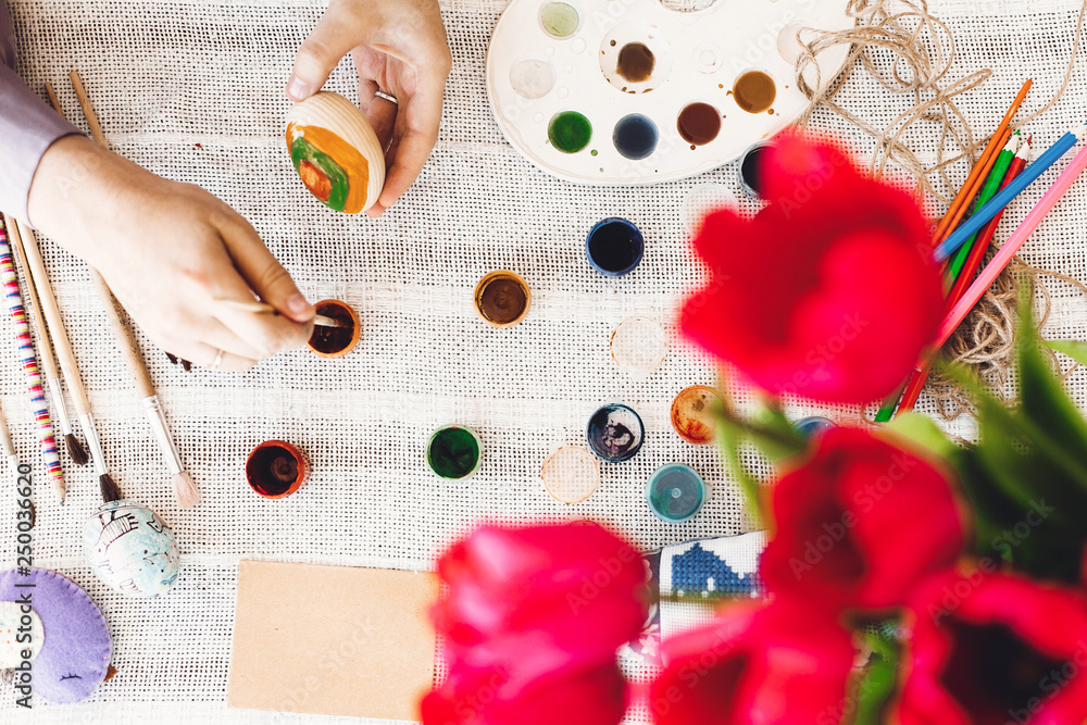 Happy Easter concept, flat lay. Hands holding and painting easter egg on rustic table with paint,brushes, tulips bouquet, space for text. Child decorating easter egg, holiday preparations Happy Easter concept, flat lay. Hands holding and painting easter egg on rustic table with paint,brushes, tulips bouquet, space for text. Child decorating easter egg, holiday preparations