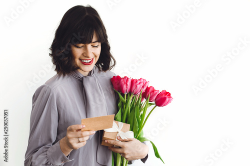 Beautiful brunette girl holding bouquet of tulips, gift box and greeting card on white background indoors, space for text. Happy young woman with flowers. Happy mothers day.