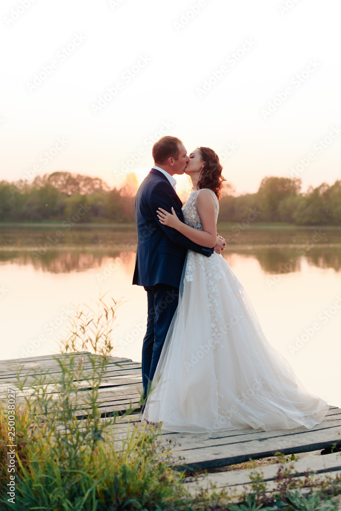 young beautiful bride and groom resting on the pier near the river at sunset