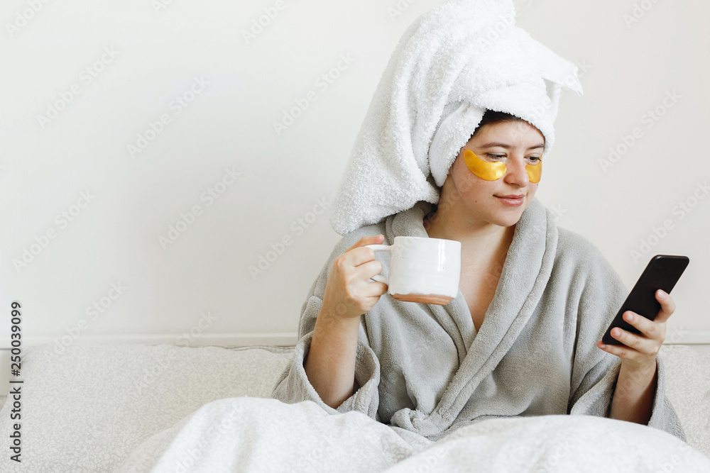 Enjoying morning concept. Happy girl drinking coffee and browsing internet. Beautiful young woman with golden eye patches in bathrobe holding cup of coffee, lying in bed and looking at phone screen Enjoying morning concept. Happy girl drinking coffee and browsing internet. Beautiful young woman with golden eye patches in bathrobe holding cup of coffee, lying in bed and looking at phone screen