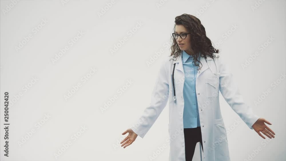 Beautiful young brunette doctor wearing medical uniform over isolated gray background. During this she has a phonendoscope on her neck, dancing and rejoicing and moving her hands. Beautiful young brunette doctor wearing medical uniform over isolated gray background. During this she has a phonendoscope on her neck, dancing and rejoicing and moving her hands.