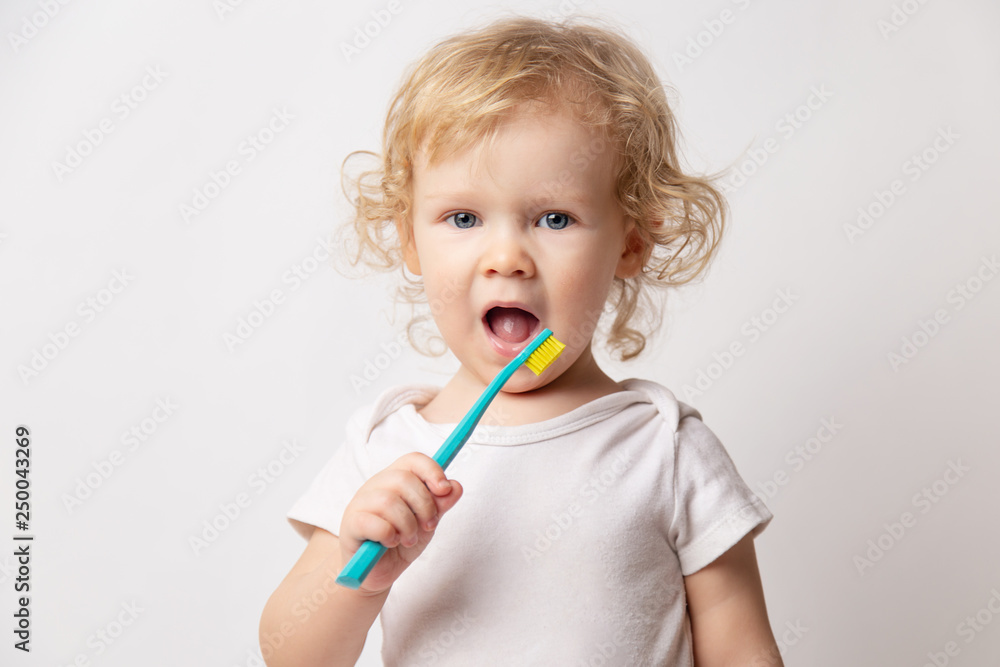 Caucasian little cute curly baby girl in white clothes body brushing her teeth with a colored blue brush on a white background. Caucasian little cute curly baby girl in white clothes body brushing her teeth with a colored blue brush on a white background.