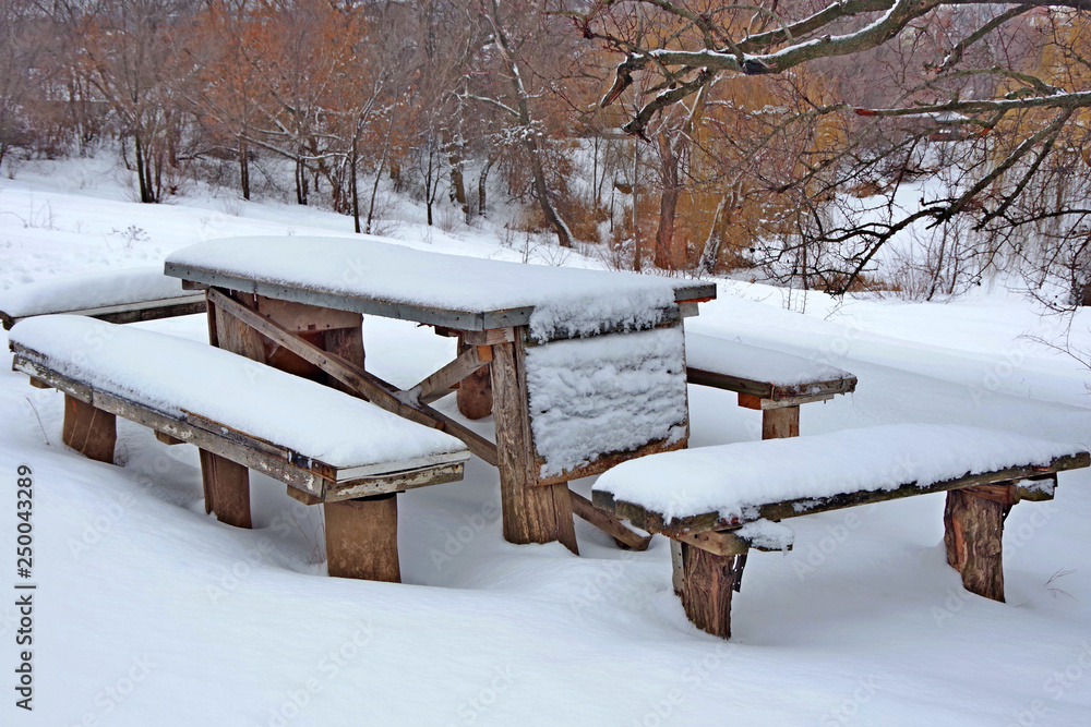 Winter. Place to rest. Cold winter day. White snow lies in a thick layer on the table and chairs. Trees and shrubs are covered with snow.