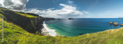 Panoramic view of the Northern Ireland shoreline. Cozy bay surrounded by the green grass covered Irish land. Overview of the white sand beach. The bright blue sky with the white clouds. Wilderness.