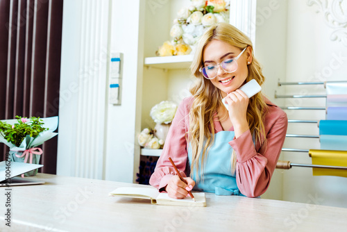 beautiful smiling female flower shop owner in glasses writing in notebook and holding smartphone