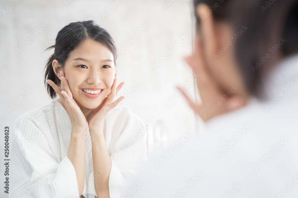 Female Skin Care. Young asian woman touching her face and looking to mirror in bathroom Female Skin Care. Young asian woman touching her face and looking to mirror in bathroom