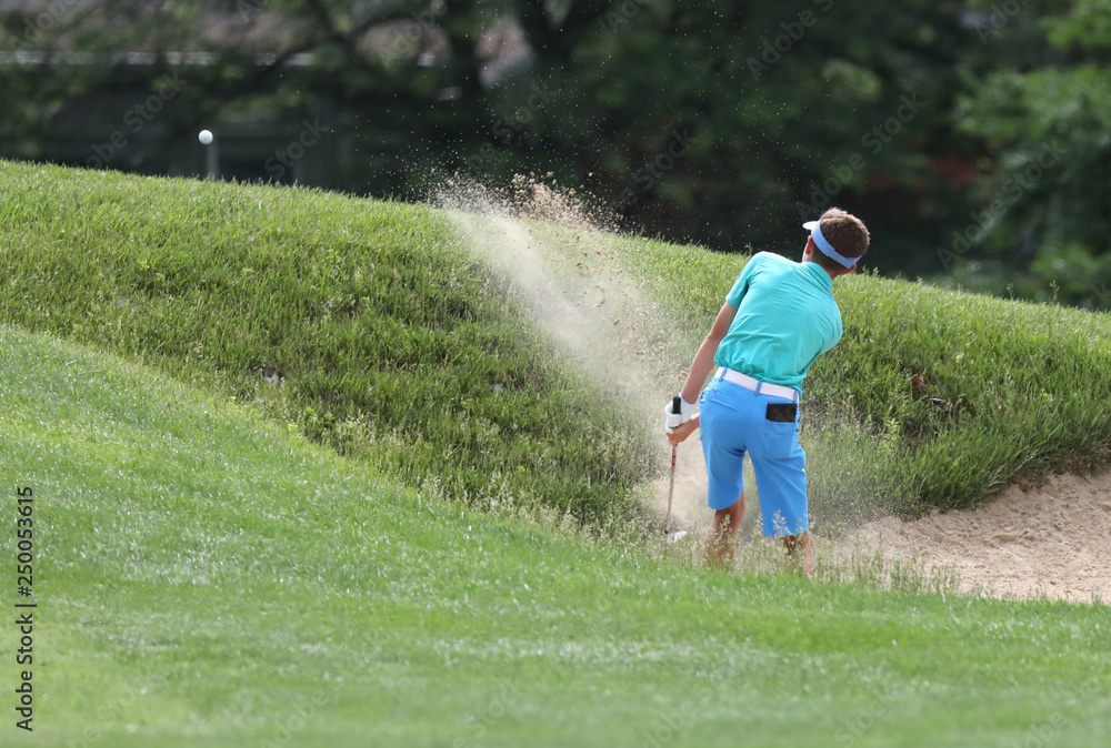a young man, competing in a golf tournament, hits a shot out of a bunker. a young man, competing in a golf tournament, hits a shot out of a bunker.