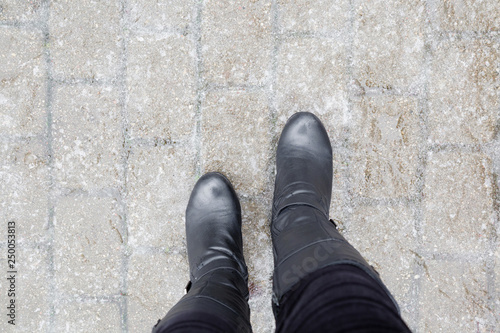 Young woman's legs in black leather boots walking on sidewalk in wet, warm winter day. Pavement covered with slippery ice. Point of view shot. Closeup. 