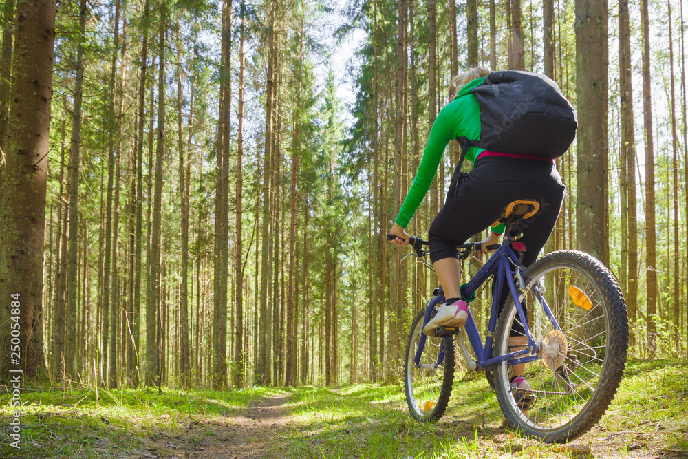 Naklejka premium Young adult woman cycling through green forest in fresh air. Backpack on back. Enjoying sport in sunny day. Outdoor activities on weekends. Active recreation in nature. Low angle view.