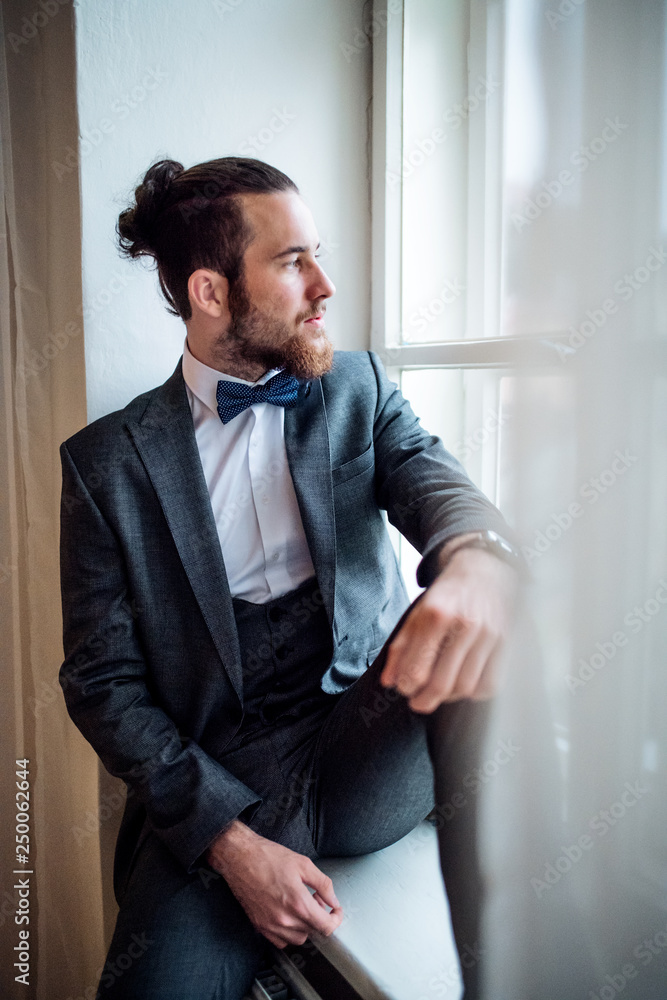 A handsome hipster young man with formal suit sitting on a window sill on an indoor party. A handsome hipster young man with formal suit sitting on a window sill on an indoor party.