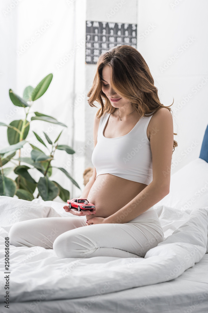 Smiling pregnant woman touching belly and holding red toy car