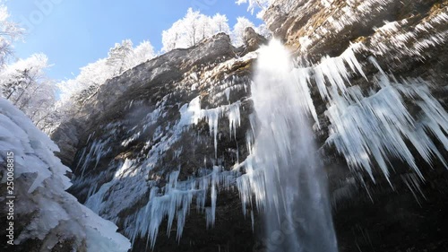 NATURE BACKGROUND, SLOW MOTION 4K Low angle wide shot of mist and ice formations forming under a waterfall falling from the side of a rocky cliff on a sunny winter day. Waterfall falling past numerous