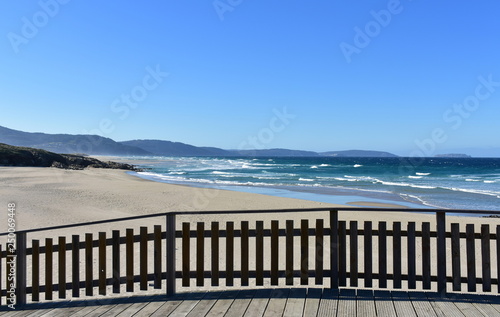Wild beach with golden sand and wooden boardwalk. Blue sea with waves and foam, sunny day. Galicia, Spain.