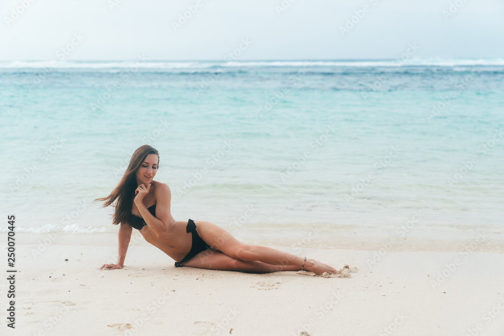 Sexy tanned girl in black swimsuit posing on sandy beach near ocean. Beautiful model sunbathes and rests