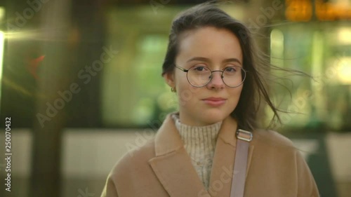 Slowmo happy gorgeous female college student brunette wearing glasses spring coat, tilting head standing square city-center lively street, train moving behind, sunlight hitting camera cover girl face