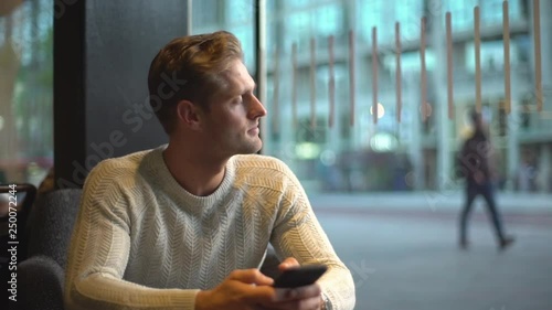 Slow motion attractive masculine european man sitting inside cafe gloomy, worried, looking outside window thoughtful space out, thinking, contemplating passersby, waiting reply phone message