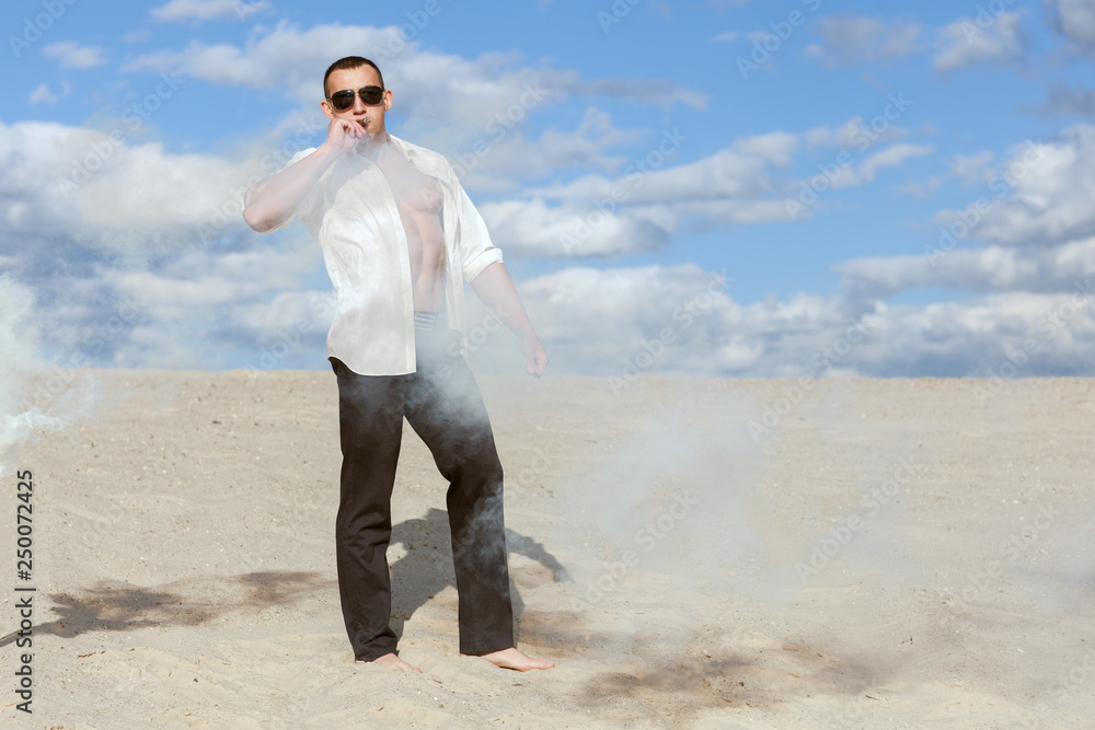 Young macho man in glasses stands amid the smoke in the desert and smokes a cigar.