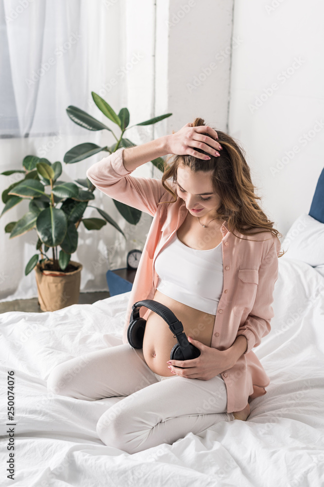 Pregnant woman sitting on bed and holding headphones on belly Pregnant woman sitting on bed and holding headphones on belly