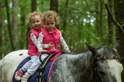 Identical twins enjoying horseback riding in the woods, young pretty girls with blond curly hair on a horse with backlit leaves behind, freedom, joy, movement, outdoor, spring, healthy lifestyle