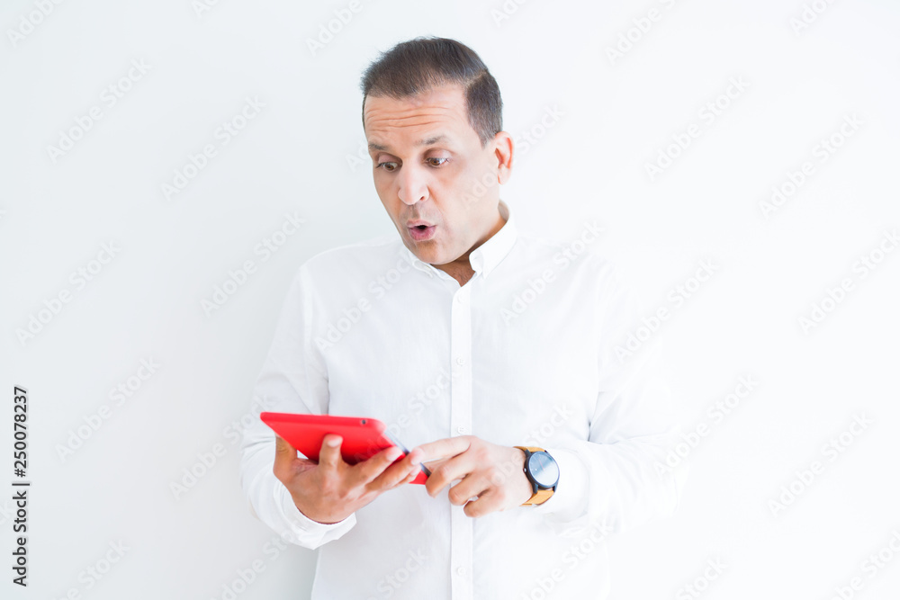 Middle age man using digital table over white wall scared in shock with a surprise face, afraid and excited with fear expression Middle age man using digital table over white wall scared in shock with a surprise face, afraid and excited with fear expression