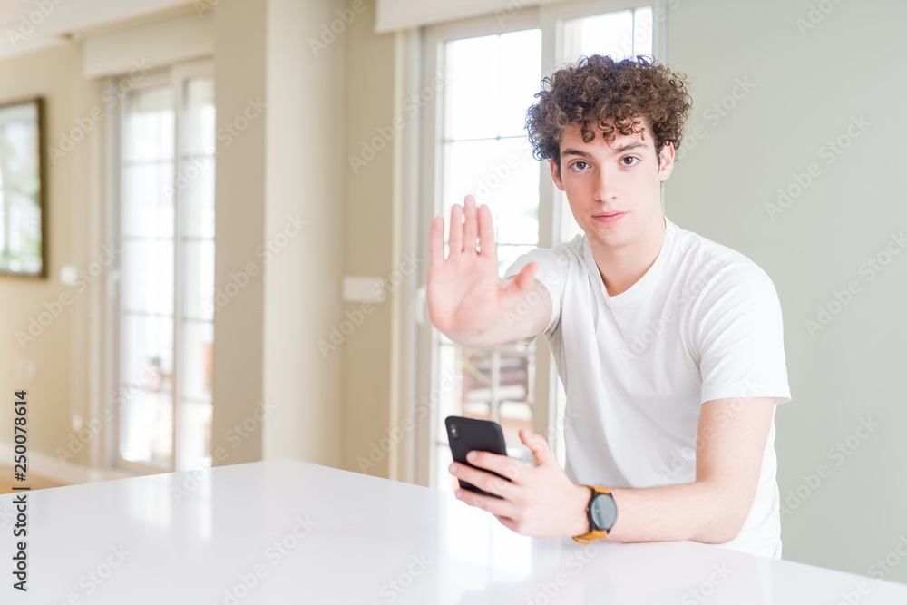 Young man using smartphone at home with open hand doing stop sign with serious and confident expression, defense gesture Young man using smartphone at home with open hand doing stop sign with serious and confident expression, defense gesture