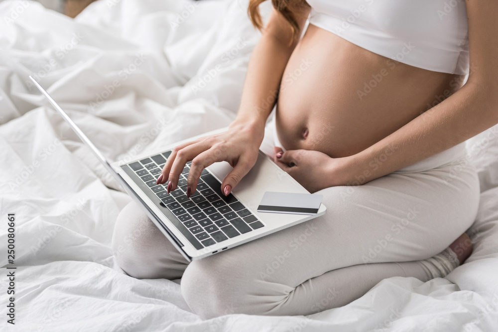 Naklejka premium Partial view of pregnant woman sitting on bed with laptop and credit card