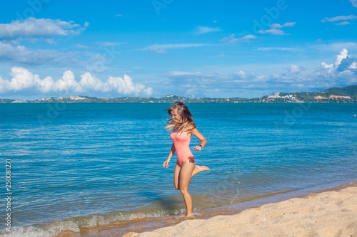 Happy young woman in pink swimsuit with long hair jumping on the tropical beach near the blue sea while sky is blue, Koh Samui, Thailand