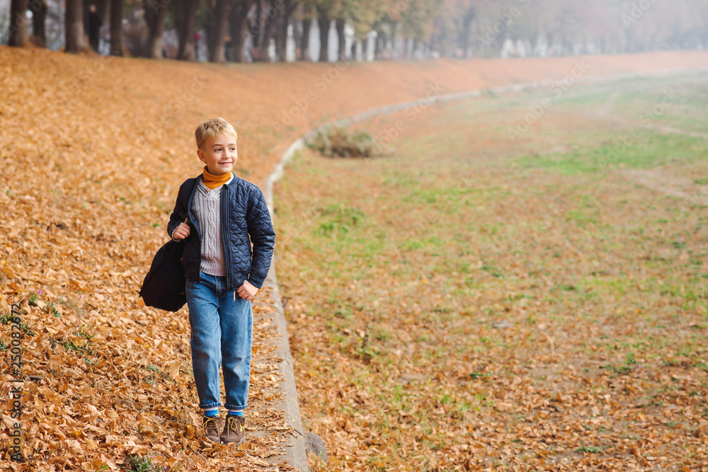Fashionable boy walking in autumn park. Stylish boy with backpack. Fashion outfit for kid. Autumn children's fashion. Cute kid enjoying fallen leaves. Happy boy at autumn day.