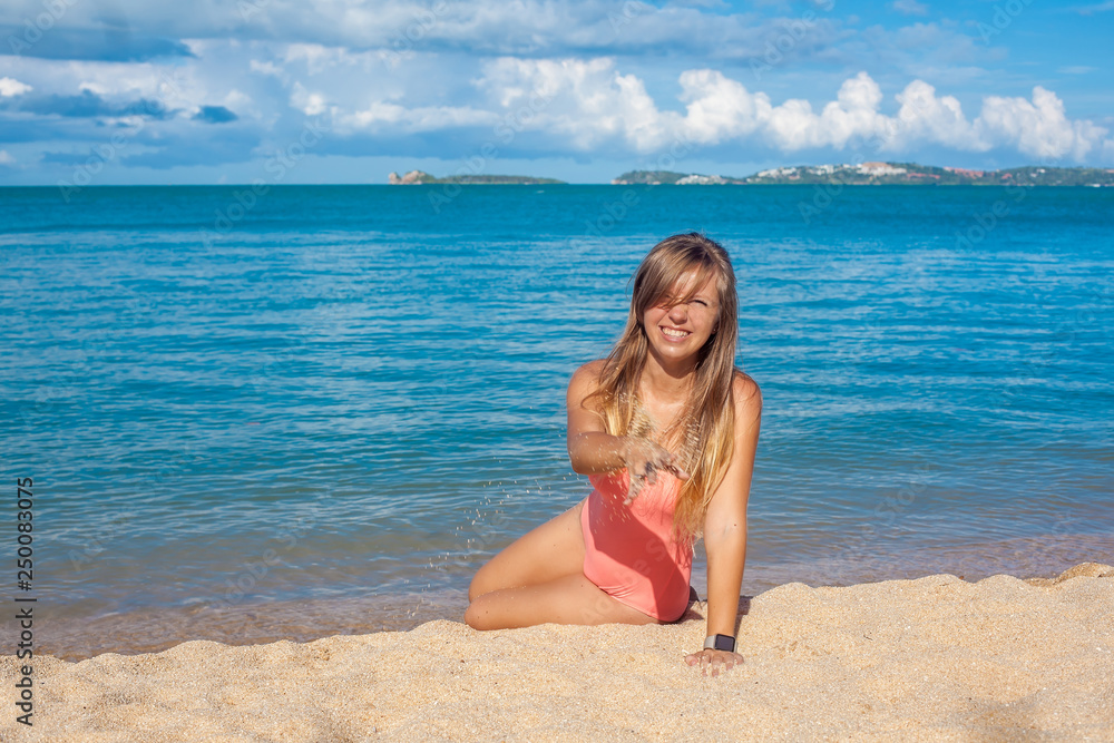 Cute smiling young woman in pink swimsuit with long hair throwing sand on the tropical beach near the blue sea while sky is blue, Koh Samui, Thailand Cute smiling young woman in pink swimsuit with long hair throwing sand on the tropical beach near the blue sea while sky is blue, Koh Samui, Thailand