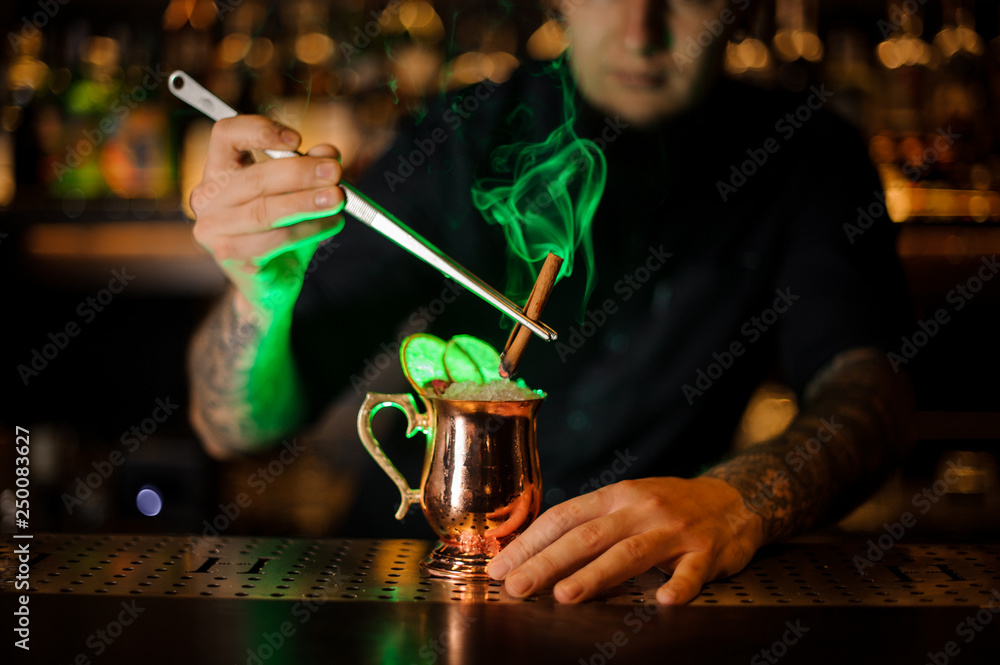 Professional bartender adding to a cocktail with dried orange a smoked cinnamon with tweezers
