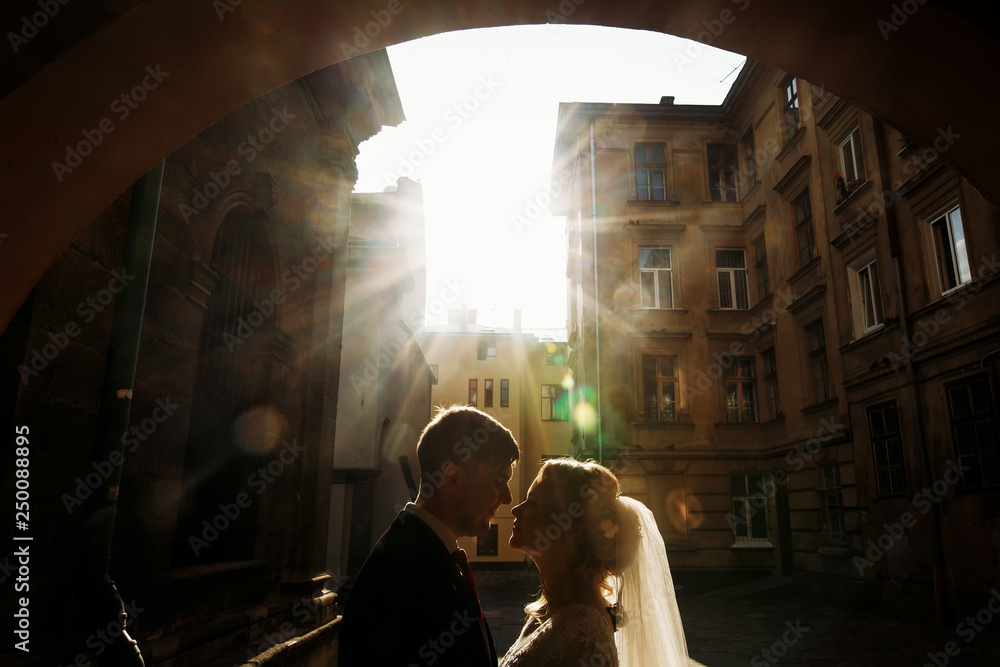 Beautiful blonde bride hugging handsome happy groom at sunset, newlywed couple kissing in old street, victorian architecture in the background