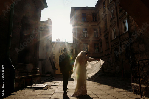 Happy couple holding hands at sunset, newlywed bride and groom posing in sunny european street, stylish husband and wife standing near old buildings