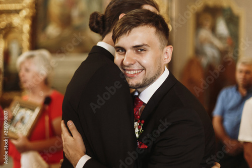 Happy, smiling groom hugging bestman after wedding ceremony in church, stylish handsome man embracing groomsman friend indoors and smiling photo