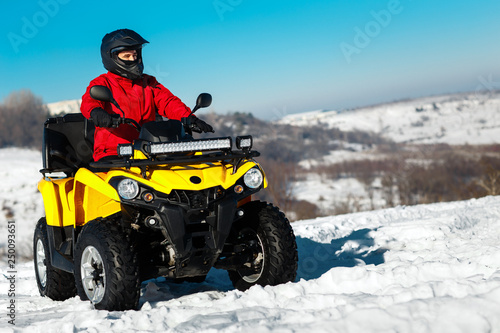 Photo of sporty young man in red warm winter clothes and black helmet on the ATV 4wd quad bike stand in heavy snow with deep wheel track. Moto winter sports.